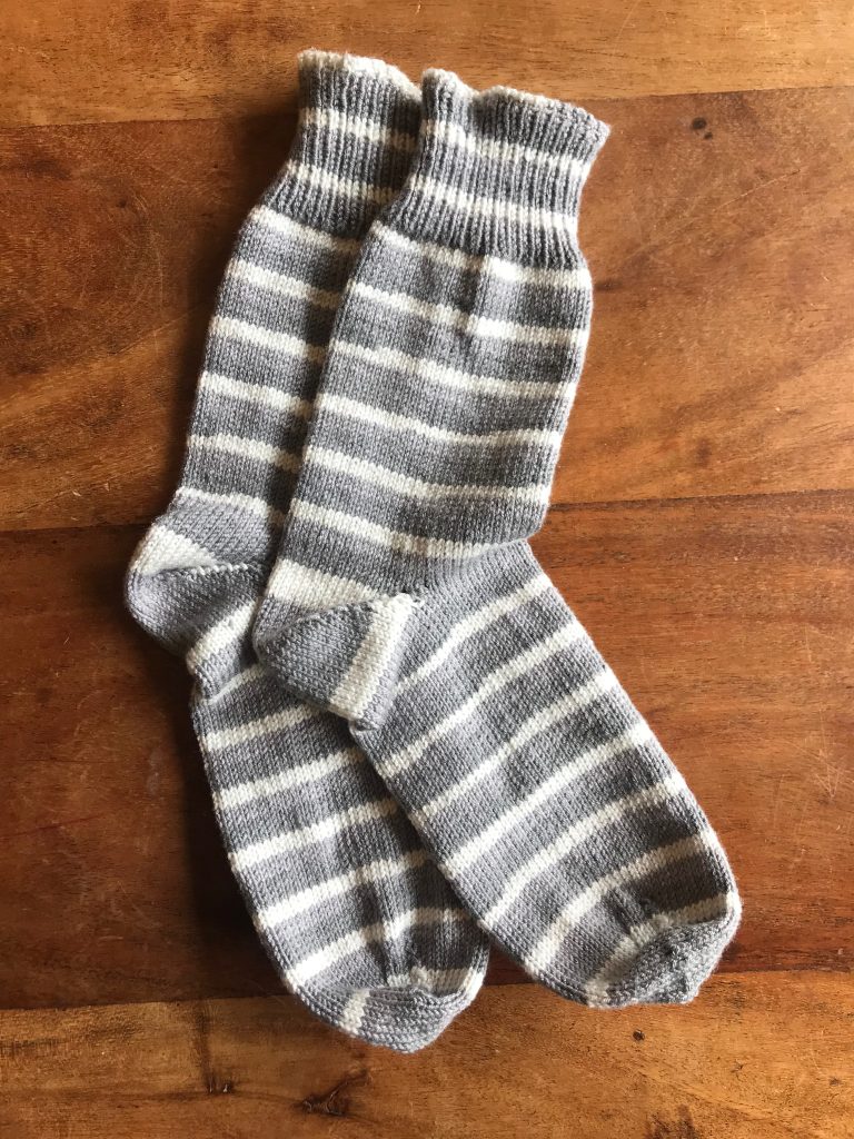 A pair of hand knitted socks on a wooden table surface. The socks are striped in two different shades of grey and white. 